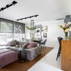 Nowoczesny salon z jadalnią i aneksem kuchennym, szara sofa, drewniany stół, kwiaty.