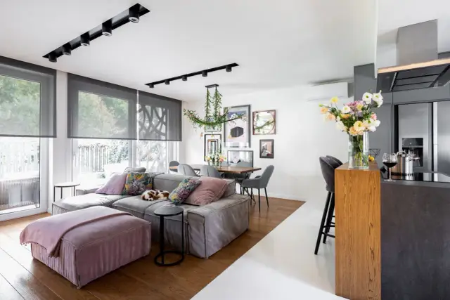 Nowoczesny salon z jadalnią i aneksem kuchennym, szara sofa, drewniany stół, kwiaty.